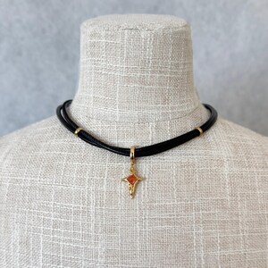 May include: A black leather choker necklace with a gold-colored pendant. The pendant is a star shape with a reddish-orange center. The necklace has gold-colored accents and is displayed on a mannequin.