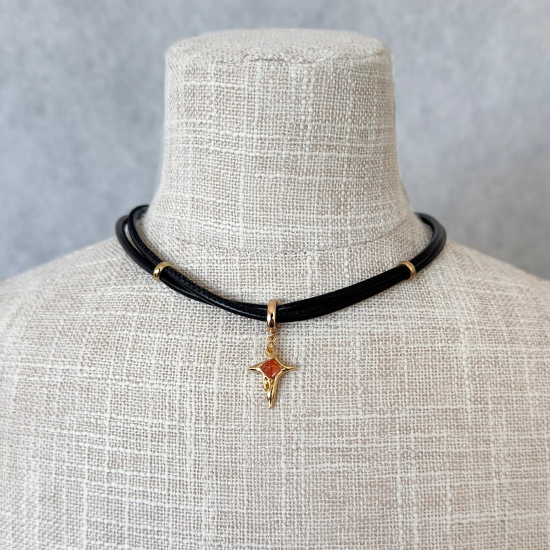 May include: A black leather choker necklace with a gold-colored pendant. The pendant is a star shape with a reddish-orange center. The necklace has gold-colored accents and is displayed on a mannequin.