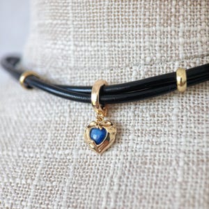 May include: A black leather choker necklace with gold accents. The necklace features a gold pendant in the shape of a heart with a blue center. The choker is displayed on a neutral fabric background.