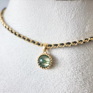 May include: A gold-coloured necklace with a round pendant featuring a green and white stone. The pendant is encircled by a twisted gold rope design. The necklace chain is a gold-coloured chain with black accents.