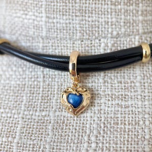 May include: A black leather choker necklace with gold accents. The necklace features a gold heart-shaped pendant with a blue center stone. The pendant is suspended from a gold ring.