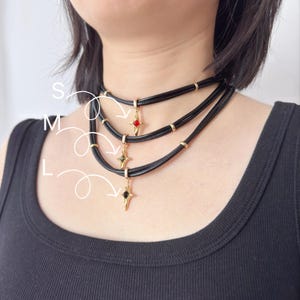 May include: Black choker necklaces with gold accents and star-shaped pendants. Each necklace features a different colored gem: red, green, and black. The necklaces are layered and worn around the neck.