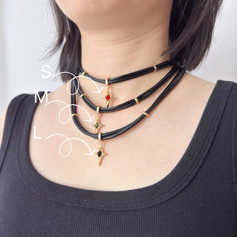May include: Black choker necklaces with gold accents and star-shaped pendants. Each necklace features a different colored gem: red, green, and black. The necklaces are layered and worn around the neck.