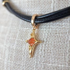 May include: A black leather choker necklace with a gold-colored star pendant. The star pendant has a central orange-colored stone. The necklace is displayed on a textured, off-white surface.