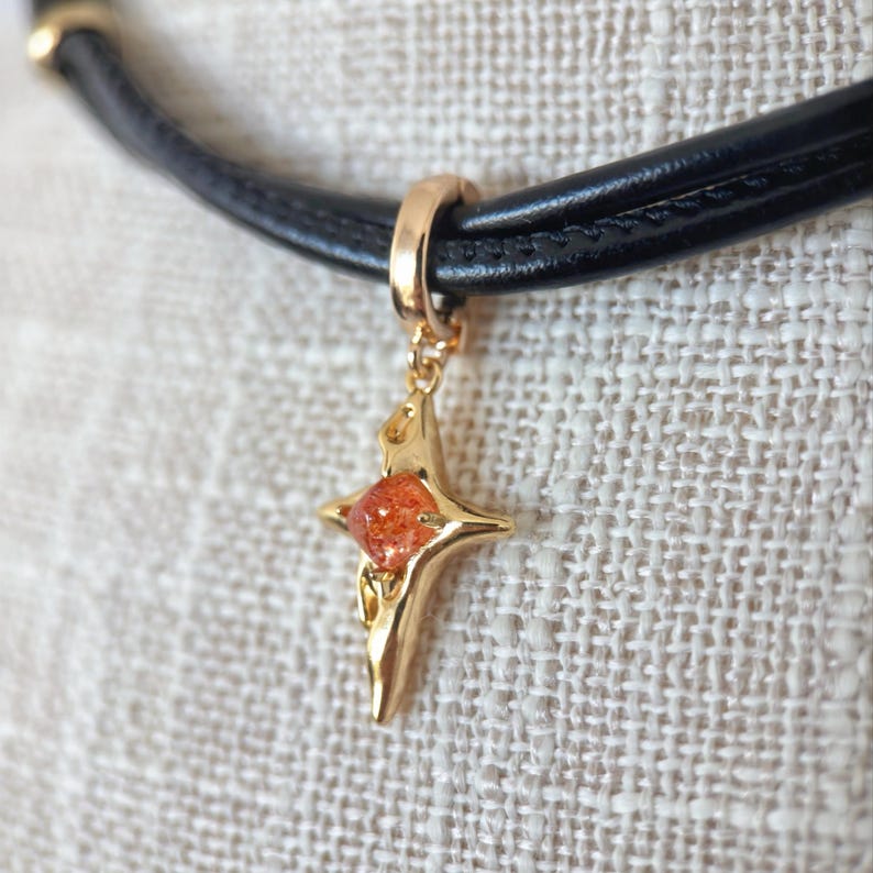 May include: A black leather choker necklace with a gold-colored star pendant. The star pendant has a central orange-colored stone. The necklace is displayed on a textured, off-white surface.
