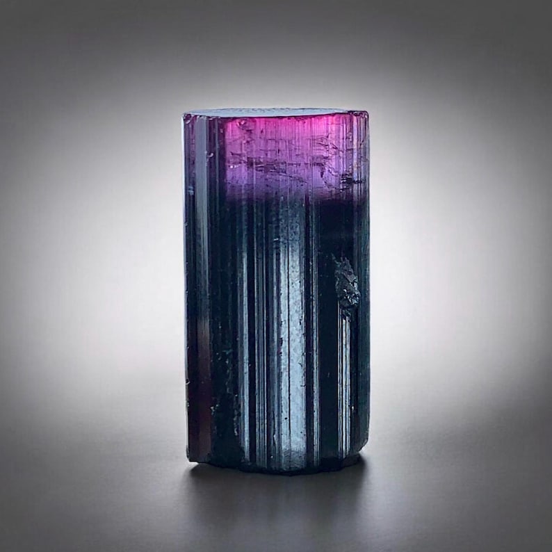 May include: A rectangular gemstone with a colour gradient. The top is pink, fading into purple, and then to a dark teal at the base. The surface has vertical striations. A mineral specimen.