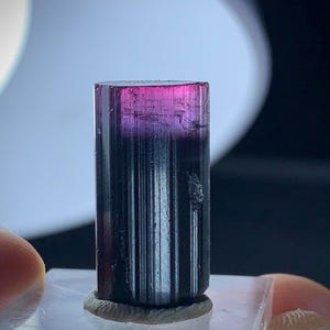 May include: A tourmaline crystal, cylindrical in shape, displaying a colour gradient from magenta at the top to a dark base. The crystal is mounted on a clear, rectangular base.