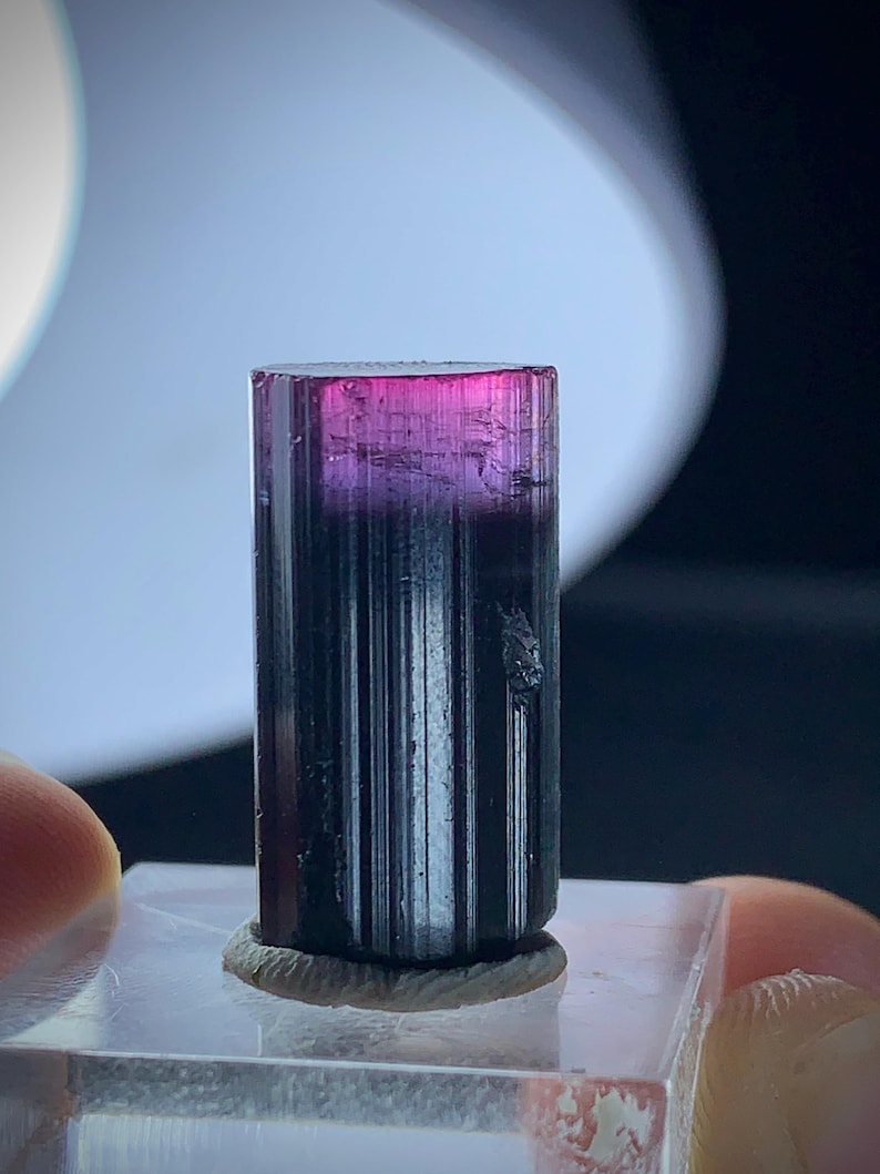 May include: A tourmaline crystal, cylindrical in shape, displaying a colour gradient from magenta at the top to a dark base. The crystal is mounted on a clear, rectangular base.