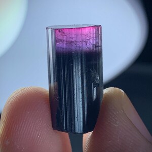 May include: A tourmaline crystal, cylindrical in shape, displaying a colour gradient. The top is a bright pink, fading into purple, then black at the base. The crystal is held between two fingers.
