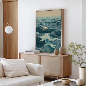 May include: A framed artwork depicts a dynamic ocean scene with waves in shades of teal, blue, and white. The artwork is displayed above a wooden cabinet with two small vases. A white sofa and a floor lamp with a spherical shade are in the foreground.
