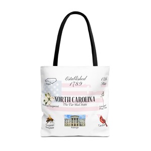 Puede incluir: Bolsa de tela blanca con asa negra que presenta un diseño del estado de Carolina del Norte. El diseño incluye el apodo del estado, "The Tar Heel State", y el año en que se fundó, 1789. La bolsa también presenta imágenes de la flor, el pájaro y el insecto del estado.