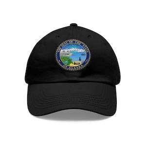 May include: Black baseball cap with a blue and gold embroidered patch featuring the seal of the state of Alaska. The seal depicts a mountain range, a ship, a moose, and a polar bear.