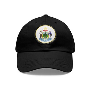Pode incluir: Boné de beisebol preto com um remendo costurado apresentando o selo do estado do Maine. O selo representa uma árvore de pinho, um fazendeiro e um marinheiro.