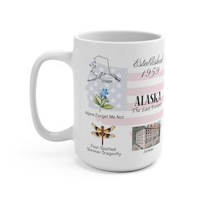 May include: White ceramic mug with a pink and blue design featuring a map of Alaska, a dragonfly, a forget-me-not flower, and the text "Established 1959 Alaska The Last Frontier Juneau".