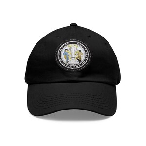 Op de afbeelding: Een zwarte baseballpet met een witte geborduurde patch met het Grote Zegel van de staat Wyoming. Het zegel toont een scène met een man die een houweel vasthoudt en een vrouw die een ploeg vasthoudt, met de woorden "GElijke RECHTEN" en "JUSTITIE" erboven.