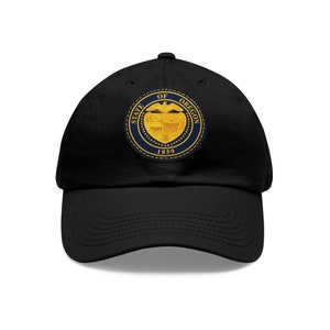 Puede incluir: Gorra de béisbol negra con un parche bordado en oro y azul que presenta el sello del estado de Oregón. El sello incluye las palabras "State of Oregon" y "1859".