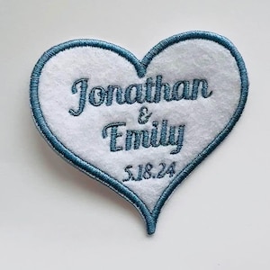 May include: A white heart-shaped patch with blue embroidery. The patch says "Jonathan & Emily 5.18.24".