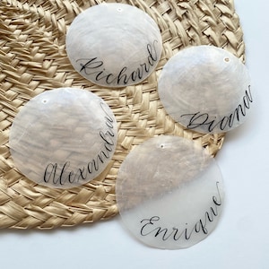 May include: Four white seashells with black calligraphy names: Richard, Diana, Alexander, and Enrique. The seashells are arranged on a woven straw mat.