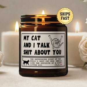 Puede incluir: Una vela encendida en un tarro de cristal marr&oacute;n con tapa negra. La etiqueta dice "MY CAT AND I TALK SHIT ABOUT YOU" con un gr&aacute;fico de gato. La vela est&aacute; rodeada de velas y flores blancas.
