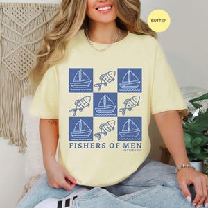以下が含まれることがあります： バター色のTシャツ。青いヨットと魚のグリッドパターン。テキスト「FISHERS OF MEN」が下に印刷されています。このシャツは快適でカジュアルなスタイルです。