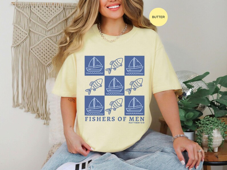 以下が含まれることがあります： バター色のTシャツ。青いヨットと魚のグリッドパターン。テキスト「FISHERS OF MEN」が下に印刷されています。このシャツは快適でカジュアルなスタイルです。