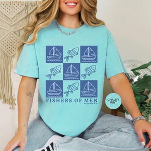 以下が含まれることがあります： 青色のTシャツ。青い正方形の中にボートと魚のグラフィックデザイン。デザインの下には「FISHERS OF MEN」の文字、その下に「Matthew 4:19」とプリントされています。シャツの色はチョーキーミントです。