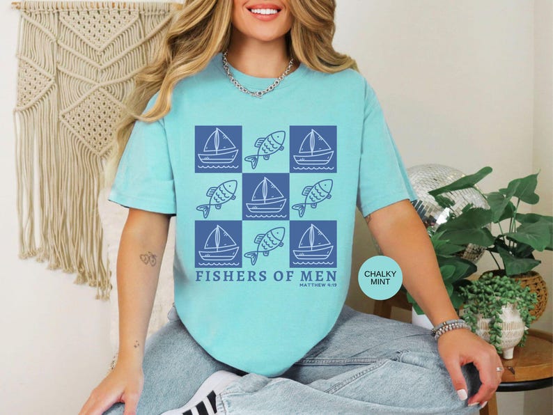 以下が含まれることがあります： 青色のTシャツ。青い正方形の中にボートと魚のグラフィックデザイン。デザインの下には「FISHERS OF MEN」の文字、その下に「Matthew 4:19」とプリントされています。シャツの色はチョーキーミントです。