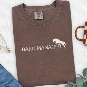 May include: Espresso-colored t-shirt with the words "BARN MANAGER" and a white horse graphic. The shirt is folded and lies next to a mug with a brown and white checkered pattern, a necklace, and blue jeans.