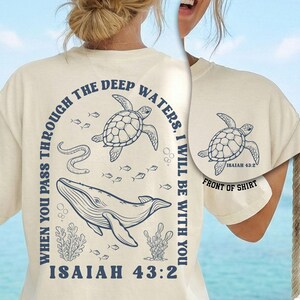 May include: Ivory-colored t-shirt featuring a marine-themed design. The back of the shirt displays a whale, turtles, and the text "WHEN YOU PASS THROUGH THE DEEP WATERS, I WILL BE WITH YOU. ISAIAH 43:2." The front of the shirt has a turtle graphic.