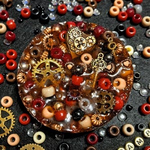 May include: A steampunk-themed resin pendant with embedded gears, beads, and a key charm. The pendant is surrounded by various beads in red, black, cream, and clear colors. Gold-colored gears and charms add detail to the design.