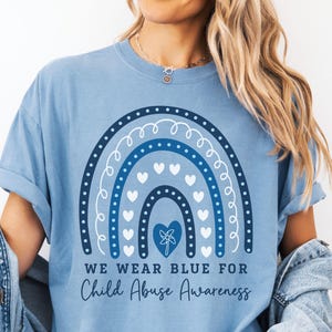 May include: Light blue t-shirt with a navy blue rainbow design, white hearts, and the text "WE WEAR BLUE FOR Child Abuse Awareness." The shirt has short sleeves and a relaxed fit. The words "Washed Denim" and "New Designs Every Week!" are also visible.