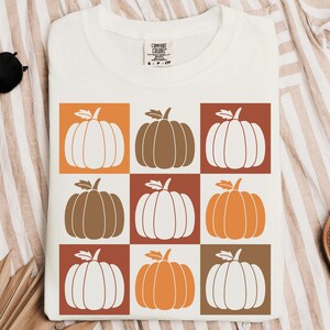 May include: White t-shirt featuring a grid pattern of pumpkins in orange, brown, and white. The pumpkins have green stems and are set against orange, brown, and white squares. The shirt is a Comfort Colors brand.