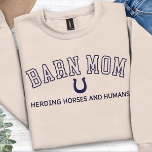 May include: A sand-colored sweatshirt with the text "BARN MOM" in navy blue, arched above a navy blue horseshoe graphic. Below, the text reads "HERDING HORSES AND HUMANS". The sweatshirt is folded, with a portion of blue jeans visible.