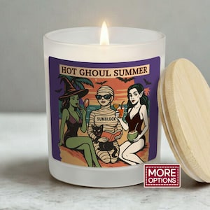 May include: A white candle with a wooden lid. The candle features a label with the text "HOT GHOUL SUMMER" and a cartoon illustration of three figures in swimwear. One figure is a witch, another is a mummy, and the third is a vampire.