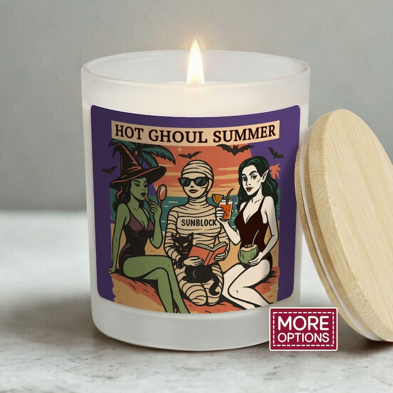 May include: A white candle with a wooden lid. The candle features a label with the text "HOT GHOUL SUMMER" and a cartoon illustration of three figures in swimwear. One figure is a witch, another is a mummy, and the third is a vampire.