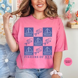 以下が含まれることがあります： 青い正方形に白いヨットと魚が描かれたデザインのピンク色のTシャツ。「FISHERS OF MEN」の文字が下にプリントされています。柔らかい素材で作られたシャツです。