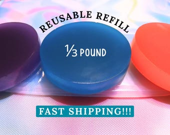 May include: Three circular, translucent wax-like discs in purple, blue, and orange. The blue disc is centered and features the text REUSABLE REFILL and 1/3 POUND. The discs are arranged on a light-colored, iridescent, textured plastic pouch.