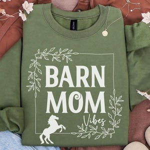 May include: A moss green sweatshirt with the words "BARN MOM Vibes" in white, framed by a leafy design and a white horse silhouette. The sweatshirt is folded on a brown surface with decorative leaves.