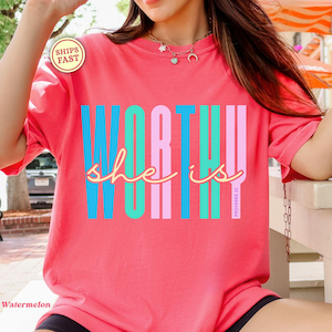 May include: A coral-colored t-shirt featuring the text WORTHY in large, vertical, multi-colored block letters in blue, teal, and pink. Overlaid in cursive script is the text she is, with the small vertical text PROVERBS 31 on the right side.