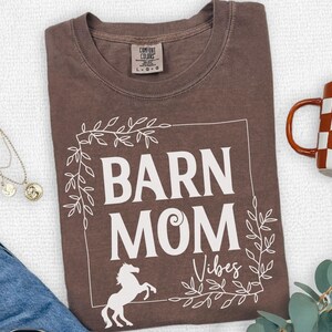 May include: Espresso-colored t-shirt with the words "BARN MOM Vibes" in white, framed by a white floral design. A white horse graphic is at the bottom. A checkered mug and jewelry are also in the image.