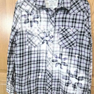 May include: A long-sleeve button-down shirt in a black, white, and purple plaid pattern. The shirt features two chest pockets with flaps, a collar, and white paint splatter accents with large flower-shaped designs. The label reads Canyon River Blues.