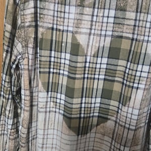 May include: A long-sleeve button-down shirt featuring a plaid pattern in shades of olive green, tan, white, and black. The fabric displays a bleached effect with a large, heart-shaped design centered on the back, created by the lighter bleached area.