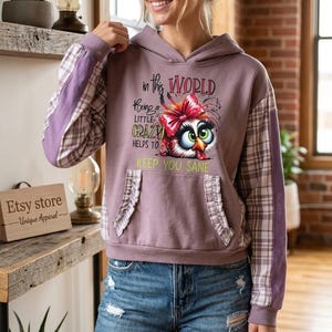 Puede incluir: Una sudadera con capucha malva de manga larga con un patrón de cuadros blanco y malva. El frente muestra un gráfico de un búho con un lazo rojo y el texto: In this WORLD Being a LITTLE CRAZY HELPS TO KEEP YOU SANE. Incluye bolsillos con volantes.