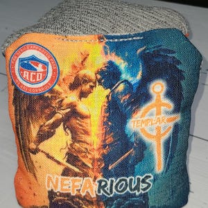 May include: A pair of cornhole bags with a graphic of two figures, one in orange and yellow flames and the other in blue and green water. The text "NEFARIOUS" is printed on the bottom of the bags. The bags are made of a grey fabric.