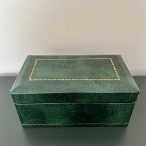 Puede incluir: Una caja rectangular verde bosque con un borde dorado alrededor de la tapa. La caja tiene un aspecto texturizado y una tapa con bisagras. La caja está sobre una superficie oscura.