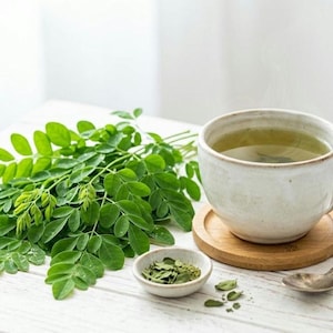 Può includere: Una tazza di tè verde è appoggiata su un sottobicchiere di legno, accanto a una piccola ciotola di foglie essiccate e un mazzetto di foglie verdi fresche. Un cucchiaio d'argento è appoggiato sul tavolo. Il tè è di colore verde chiaro.