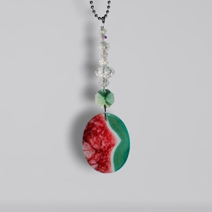 May include: A decorative hanging ornament featuring a red and green oval-shaped stone pendant. The pendant is suspended from a black chain and adorned with clear crystal beads and a faceted green crystal. The stone has a unique, natural pattern.