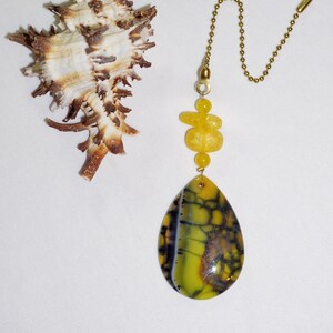 Op de afbeelding: Een decoratieve trekketting voor een plafondventilator met een geel en zwart druppelvormige hanger. De hanger is verbonden met een messing ketting en gele kralen. Een getextureerde zeeschelp staat ook in de afbeelding.