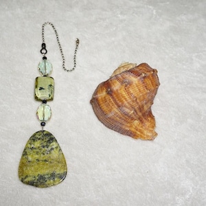 May include: A decorative ceiling fan pull chain with a green and black stone pendant and a seashell. The pull chain has a silver chain and beads in shades of green and clear. The seashell is brown and tan.