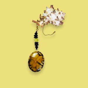 Könnte beinhalten: Ein Anhänger aus braunem und gelbem Achat mit schwarzen und grünen Perlen, die an einer goldenen Kette hängen. Der Anhänger ist an einer Muschel befestigt.